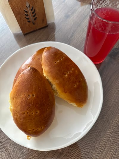 A white plate holds three golden-brown baked goods with small holes poked into their surface. To the right of the plate, a clear glass contains a vibrant red liquid, likely a beverage. In the background, a wooden napkin holder with a carved leaf design sits behind a stack of white napkins. The objects are arranged on a dark wooden surface, possibly a table, which appears to be in the city of Znamya Oktyabrya, Russia. The scene is likely indoors, given the lighting and presence of household items.