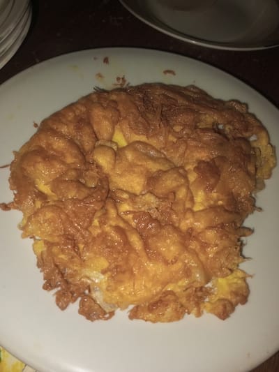 An overhead shot reveals a single, deep-fried egg dish, golden to dark brown and irregularly shaped, presented on a white, round plate. The dish displays a craggy, crispy texture with visible oil residue. Partially visible in the background are a stack of white plates on the upper left and the rim of another white plate on the upper right, resting on a dark surface. The ambient lighting is low. Location: Doramamidi, India.
