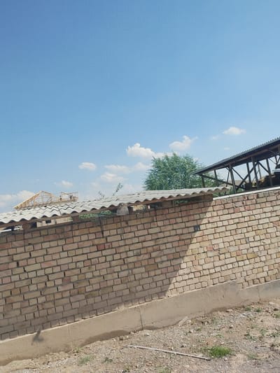 The image shows an outdoor scene under a clear blue sky with a few scattered white clouds. The primary subject is a brick wall constructed from light-colored bricks, with a concrete foundation visible at its base. The wall extends across the lower portion of the frame.
Above the brick wall, there are several structures with wavy, corrugated roofs, possibly made of asbestos or similar material. In the background, a wooden structure with a metal roof is visible, appearing to be a shelter or open-air building, with hay bales or similar material stacked inside. Further in the distance, a lush green tree stands out against the sky.
On the roof of the wooden structure, a few people are visible, seemingly engaged in some activity, possibly construction or maintenance. Another structure with a wooden frame and roof is under construction, with wooden beams and supports clearly visible.
The lighting suggests it is daytime, likely late morning or early afternoon, given the bright sunlight and the distinct shadows cast by the structures. The weather appears to be fair and warm. The scene appears to be in a rural or semi-rural area, possibly a farm or a construction site. There is no visible text in the image. The overall mood is one of outdoor activity and construction in a sunny environment.