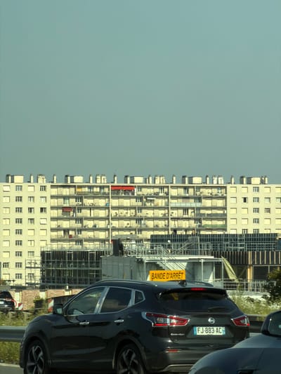 The image shows a view from a highway in Vitry-sur-Seine, France, with a large apartment building dominating the background. The apartment building is light beige with many balconies, some with red or yellow awnings. Various windows are visible, some open and some closed. The roofline of the building features multiple chimney-like structures. In front of the apartment building, there are ongoing construction or renovation activities, with scaffolding, white modular structures, and industrial fencing visible. A yellow traffic sign with the text "BANDE D'ARRET" (shoulder) is positioned on the right side of the highway. In the foreground, a dark-colored Nissan Qashqai SUV is driving on the highway, displaying the license plate "FJ-883-AC". The sky is a clear, pale blue-grey.