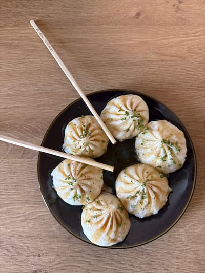 The image shows a top-down view of a dark plate filled with six steamed dumplings, likely soup dumplings (xiaolongbao), arranged on a light brown wooden table. The dumplings are plump and white, with visible pleats at the top, suggesting they are freshly prepared. Each dumpling is drizzled with a light brown sauce and garnished with finely chopped green herbs, likely chives, and flecks of black pepper.
Two light-colored wooden chopsticks are placed diagonally across the plate, with their tips resting on two of the dumplings, indicating that the food is ready to be eaten. One of the chopsticks has visible text printed on it in what appears to be Asian characters and some small logos.
The lighting is bright, suggesting it's daytime, and the scene is set indoors on a wooden surface. The overall impression is of a well-presented dish, ready to be enjoyed. The location context provided places this scene in Damme, Belgium. No people are visible in the image.