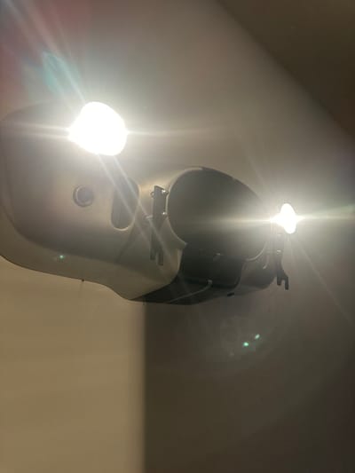 A white, sculpted object with dark interior elements hangs on a wall. It's illuminated by two bright light sources: one near the top and another towards the bottom, creating intense glare and light streaks. The object seems to have some mechanical parts and an oval-shaped mesh opening on its lower front surface. The wall appears to be painted in a light shade.