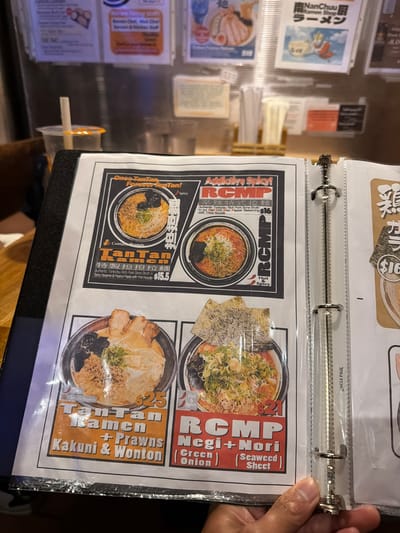 The image is a close-up shot of a menu within a binder, likely in a restaurant. The menu features several ramen dishes, with vibrant photos and descriptions. The binder has metal rings, suggesting it's a professional menu. The overall lighting is bright, indicating it's daytime, and the setting appears to be indoors.

The menu items are described in both English and Japanese, with the following visible text: "NanChuu Ramen Shop", "RCMP", "TanTan Ramen", "Kakuni & Wonton", "Negi+Nori (Seaweed) (Green Onion)", and prices like "$15.5", "$16", and "$25". Some descriptions mention "Authentic Tonkotsu Rich Pork Bone Broth", "Spicy Sesame & Peanut Paste", and "Red Chili Miso Pepper Seasoning". Some dishes are explicitly labeled "Hot & Spicy" and one indicates "Contains Peanuts". A small section of another menu page is visible at the top, with text including "$16".

The presence of a hand holding the binder open suggests someone is either viewing the menu or taking a picture of it. The style of the menu and the dishes advertised, along with the "Richmond, Canada" context, point towards a Japanese ramen restaurant. The emotions conveyed are neutral, focusing on presenting the food items.