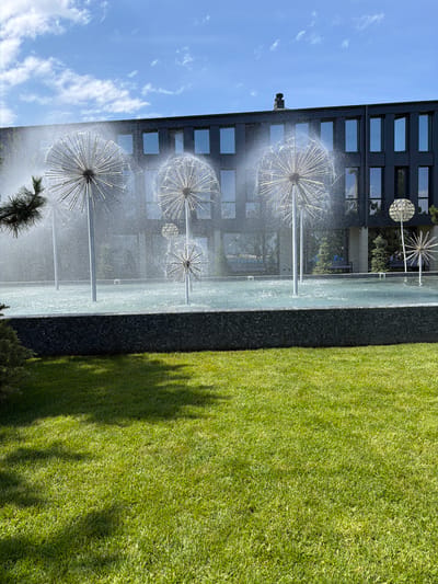 The image depicts a modern architectural setting with a water fountain feature. In the foreground, a meticulously manicured green lawn is visible, with shadows from unseen trees cast across it. Behind the lawn is a dark tiled retaining wall surrounding a rectangular pool of water. The pool contains several large, spherical metal sculptures resembling dandelion seed heads, from which water sprays upwards, creating a mist. These sculptures are mounted on tall, slender poles. In the background, a contemporary, dark-colored building with a dark facade and numerous evenly spaced rectangular windows is prominent. The sky above is blue with scattered white clouds. The scene appears to be a public space or a park area adjacent to the building. The provided information indicates the location is Khotianivka, Ukraine. There are no people visible in the image.
