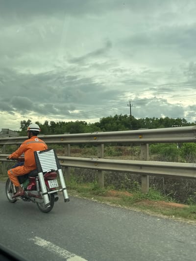 An individual wearing an orange uniform and a white helmet is riding a red motorcycle on a paved road in Hải Lăng Commune, Vietnam. The motorcycle is equipped with a silver ladder secured to the rear. The background features a guardrail, trees, utility poles, and a cloudy sky. The overall scene depicts a person commuting with equipment in a rural or semi-urban setting.