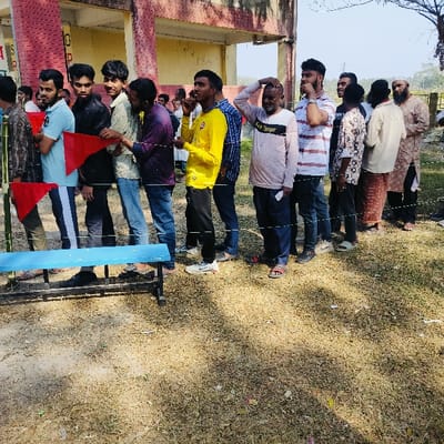 A line of men is waiting outdoors, possibly to vote, indicated by red flags. Some individuals are on their phones, others are adjusting their attire, and one man scratches his head. They are dressed in casual clothing, including jeans, t-shirts, and traditional attire. A blue bench stands nearby. The background shows a building and trees under a clear sky.