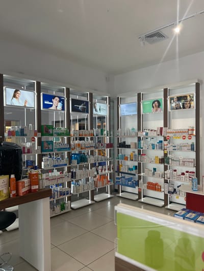 The image shows a pharmacy located at 9.94 latitude and -84.156 longitude in San Rafael, Costa Rica. The pharmacy has several rows of shelves with various personal care products and a counter with a cash register in the foreground. There are several small flat-screen monitors displaying advertisements for various products. The pharmacy is clean and well-lit with white walls and a tiled floor. There is a white coffee maker on a counter in the foreground. The pharmacy is empty of people.