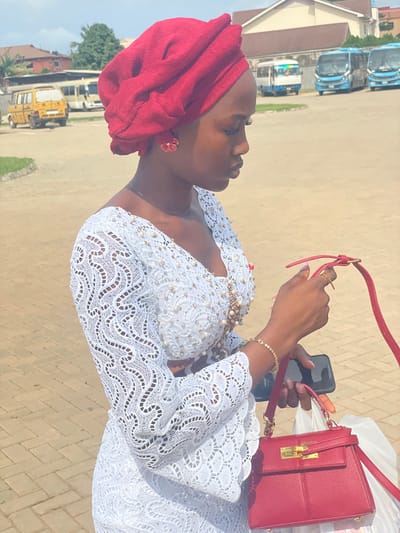 A woman in Otta, Nigeria, stands in an outdoor paved area, possibly a parking lot or courtyard, holding a smartphone in her right hand. She wears a white lace dress adorned with pearls and a red head wrap. She also has a gold bracelet on her right wrist. A red purse with a gold clasp is held in her left hand. The backdrop includes buildings, parked buses, and other vehicles.
