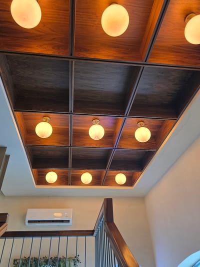 A low-angle interior shot depicts a wall with a large wooden shelving unit featuring illuminated spherical light fixtures. The shelving unit is composed of dark wood, with some sections holding light fixtures and others left empty.

Adjacent to the shelving unit, a wall-mounted air conditioning unit is visible. To the right, a wooden structure partially obscures a view of a window with green foliage visible outside.

The location is likely Tijuana, Mexico, but this information is not directly verifiable from the image content. No people are visible.