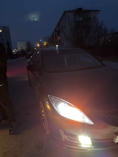 The image depicts a dark urban scene, likely at dusk or night in Zhatay, Russia. Dominating the foreground is a black car with its headlights illuminated. The car's front is angled towards the viewer.

A portion of a person is visible in the lower part of the frame, wearing a dark jacket with a white Nike swoosh, and black and white sneakers.

The backdrop consists of buildings, the structures are unlit and silhouetted. A few street lights are visible in the mid-ground, casting a faint glow on the street. The overall environment appears to be a residential street with limited illumination.