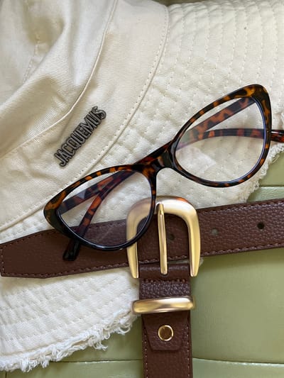 An overhead, close-up shot shows a tortoise-shell patterned pair of glasses, a brown leather belt with a gold buckle, and a cream-colored bucket hat with a silver "JACQUEMUS" logo. The items are arranged on a light green surface, possibly a sofa or a piece of furniture. The hat has frayed edges, suggesting it might be a denim or canvas material. The belt is textured and has a classic rectangular buckle. The glasses are cat-eye shaped and have a bold, thick frame. The lighting is bright and appears to be natural daylight. The scene is static and there are no people visible. The image does not provide any information to suggest it is located in Starobaltachevo, Russia.