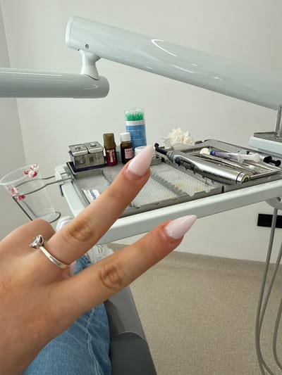 The image shows a close-up view of a person's hands in what appears to be a dental or medical setting. The subjects are the person's hands, with long, light pink, almond-shaped acrylic nails. A silver ring with a prominent diamond is visible on the ring finger. The hands are positioned in front of a tray of dental instruments.
Objects visible include various dental tools in a metal tray, such as what looks like a dental probe, a syringe, and other small instruments. There are also several small vials of what could be dental materials, some with colored liquid. A white cloth or gauze is also present. In the foreground, a clear plastic cup containing small red fragments is suspended by metal clips. To the left, a large, white, articulated dental light fixture is partially visible. Blue jeans are visible in the background, suggesting the person is seated.
The scene is brightly lit, suggesting an indoor environment, likely a dental clinic given the instruments. The overall impression is one of clinical readiness, though the prominent nail enhancements and ring suggest a personal touch within the professional setting. There is no visible indication of the time of day or weather. The location cues are not specific enough to pinpoint Strumica, North Macedonia, based solely on the visual information. No specific emotions are conveyed, beyond the neutral presentation of the hands and equipment. No legible text is present in the image.