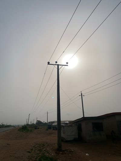 The image captures an outdoor scene dominated by utility poles and power lines against a bright, hazy sky. The overall impression is one of strong backlighting.
In the upper-left quadrant of the frame, a brilliant, almost perfectly circular and intensely white light source, presumed to be the sun, shines through a thick haze. The sky around it is a very light gray or off-white, indicating either heavy cloud cover or atmospheric haze, which diffuses the sunlight.
Several utility poles are visible. A large, dark-colored pole stands prominently in the mid-left portion of the image, extending from near the bottom edge upwards. It is seen in silhouette against the bright sky, clearly showing insulators and cross-arms from which multiple power lines extend. These thin, dark power lines crisscross the upper and middle sections of the sky, running horizontally across the frame and connecting to other poles. Another utility pole is situated further back and to the right, and more poles can be seen receding into the distance along a path.
On the far right side of the image, a structure, likely a building, is partially visible. It appears dark, possibly due to being unpainted or in shadow, with a rough texture. A corrugated metal roof is distinguishable at the top edge of the building. There's a dark, rectangular opening that might be a window or doorway. Below this main structure, a lighter-colored, possibly temporary shelter or stall, perhaps covered with a tarp, is also present.
The ground along the right side of the image is a dirt road or path, uneven and appearing somewhat barren. Sparse patches of green vegetation or grass can be seen along the edge of the path, particularly further into the background and on the extreme right. Some debris is scattered on the ground near the building. In the distance, vague shapes of what might be vehicles or other structures are discernible but indistinct due to the distance and atmospheric conditions. A faint blue-green lens flare is visible in the middle-right area of the sky, consistent with the camera pointing directly at the sun.
The intense brightness of the sun and the hazy conditions suggest it could be midday or early afternoon. There is no visible text in the image.