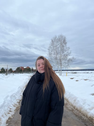 The image depicts a female individual standing outdoors in a snowy landscape, identified as Kalinino, Russia. The subject, an adult female with long brown hair, is positioned slightly off-center to the right, looking directly at the camera with a neutral expression. She wears a black, high-collar, puffy winter jacket.
In the foreground, a path, possibly dirt or gravel, is partially covered by snow, with snowbanks lining its edges. To the left of the path, in the midground, several residential structures with orange-red roofs are visible amidst evergreen trees and a fence line. Utility poles with overhead lines are also present. To the right of the path, a bare deciduous tree, appearing to be a birch, stands prominently. Beyond the tree and extending to the right, a wide, snow-covered field is visible, showing patches of exposed earth. A dark tree line forms the distant horizon. The sky is overcast with multiple layers of gray clouds. The overall scene suggests a cold, possibly late winter or early spring day.