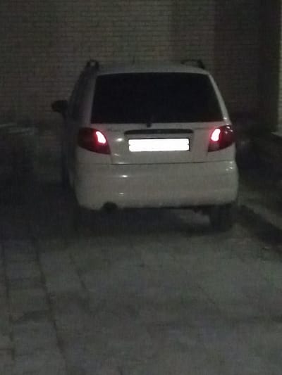 A white hatchback car is parked in a dimly lit, possibly indoor, parking area. The rear of the vehicle is visible, showing the rear window, tail lights, bumper, and license plate area. The car's tail lights are illuminated. The surroundings appear to be a brick wall on the left and a dark, possibly concrete or paved, floor. The overall lighting suggests it is nighttime.