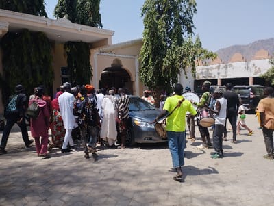 The image depicts a group of approximately twenty-five people gathered in front of a building in Kaltungo, Nigeria. Located at the coordinates 9.816 latitude, 11.315 longitude, the scene includes several individuals playing drums, a gray car parked in front of the building, and several other unidentified people standing around the car and drummers. Most individuals are dressed in casual clothing. The building is a two-story structure with a tiled roof and a large courtyard in front. The courtyard is paved with grey concrete tiles and is bordered by a green lawn.  A large tree is visible on the left side of the building. There are several other buildings visible in the background, including a multi-story building with a white facade and a large, flat roof.