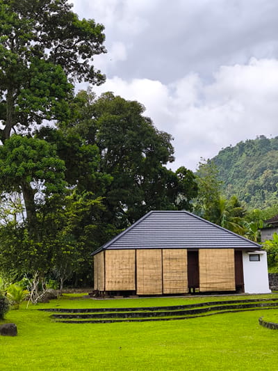 The photograph captures a building in Sidemen, Indonesia. The building has a gray-tiled roof and bamboo panel siding. It is framed by lush greenery, including large trees to the left and forested hills in the background. The foreground consists of a well-maintained green lawn with terraced stone steps. The sky is overcast.
