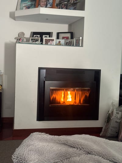 An interior scene in Nogueira e Silva Escura, Portugal, depicts a residential living space. A dark-framed, built-in fireplace insert is active, showing prominent orange and yellow flames behind its glass panel. Above the fireplace, two white floating L-shaped shelves are mounted on a white wall. The lower shelf displays multiple framed photographs, primarily depicting infants and children, and a silver-colored gingerbread-man-shaped figurine on the left. The upper shelf also contains various framed photographs, including one with a visible boat image. A dark brown wooden baseboard runs along the bottom of the white wall. On the floor in the foreground, a light grey, textured, fluffy blanket is partially visible. To the right of the fireplace, a translucent plastic bag containing dark wood pellets rests against the wall on a grey carpeted surface, adjacent to a visible dark wooden floor section. No individuals or direct interactions are depicted.