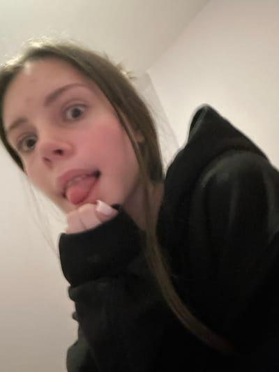 A girl in a black hoodie tilts her face towards the camera, sticking her tongue out playfully. Her light brown hair frames her face. Her eyes are wide and light brown. The background appears to be a white wall. The hoodie's sleeve and hand are visible near her face. The low angle creates a somewhat distorted perspective.