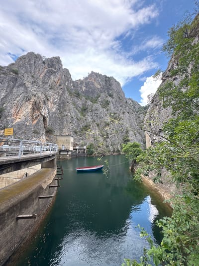 This is a photograph of the Matka Dam and Lake in Dolna Matka, North Macedonia. The image captures a view of the dam's concrete structure and a body of dark green water. A small, red and white boat is moored in the lake. Steep, rocky canyon walls rise on either side of the water, with sparse vegetation clinging to the slopes. The sky above is partly cloudy with a mix of white clouds and blue sky. A small building, likely part of the dam's infrastructure, is visible on the right bank. The foreground shows the textured concrete of the dam wall.