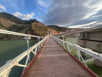 A wide-angle, full outdoor shot depicts the Ait Mazigh suspension bridge in Morocco, featuring a wooden walkway and white, truss-style railings, spanning a body of water. To the left, a mountain range with sparse vegetation rises, displaying reddish-brown soil and rocky outcrops. The sky is partially cloudy, with a mix of blue sky and darker, overcast areas. To the right, a parallel, concrete structure with red-and-white railings is visible. The water reflects the colors of the sky and surrounding landscape. The perspective is from the center of the wooden bridge, looking towards the tower and distant shore.