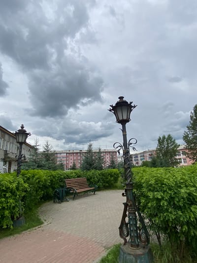 The image depicts an overcast day in a city park in Belorechensky, Russia. In the foreground, a decorative, dark metal lamppost stands beside a pathway made of light-colored paving stones with red accents. To the left of the pathway, a lush green hedge lines the area, and to the right, the hedge continues, obscuring much of the background. Behind the hedge, a wooden bench with dark metal legs sits near a trash can. Further in the background, several trees, including what appear to be coniferous trees, are visible. Beyond the trees, apartment buildings with pink and muted-colored facades and numerous windows are situated under a heavy, cloudy sky. Another lamppost, similar in design to the one in the foreground, is partially visible to the left of the apartment buildings. The overall scene suggests a quiet, urban outdoor space.