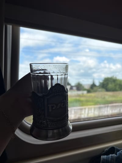 A hand holds a glass of water with condensation on the outside, resting in a metal holder with ornate carvings and Russian lettering, against a blurred background of a window on a train. Outside the window, a field with scattered trees and buildings can be seen under a partly cloudy sky. The scene is captured during daylight hours.
