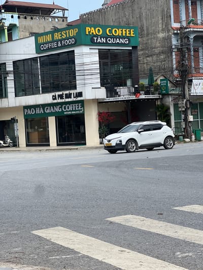 The image captures a street scene in Tan Quang Commune, Vietnam, featuring a white SUV parked on the right side of the road, partially obscuring a pedestrian crossing. The vehicle, identified by license plate "22H-037.42", is a modern crossover.

In the background, a multi-story building is prominently displayed, housing several businesses. The upper facade of this building features a large, green sign that reads "PAO COFFEE TAN QUANG", with smaller text above indicating "MINI RESTOP COFFEE & FOOD". Below this, another green sign for "PAO HA GIANG COFFEE" is visible, along with a hotline number. The lower levels of the building have large glass windows.

Adjacent to the coffee shops, there are other establishments, including one with a green sign pointing towards "COFFEE PIZZA HERE". A green awning and some potted plants are also visible. The street is paved and shows evidence of wear, with fallen leaves scattered on the asphalt and the pedestrian crossing. The sky is overcast. The scene is typical of a commercial area in a Vietnamese town, with a mix of modern businesses and older architecture, and the ubiquitous presence of electrical wires overhead.