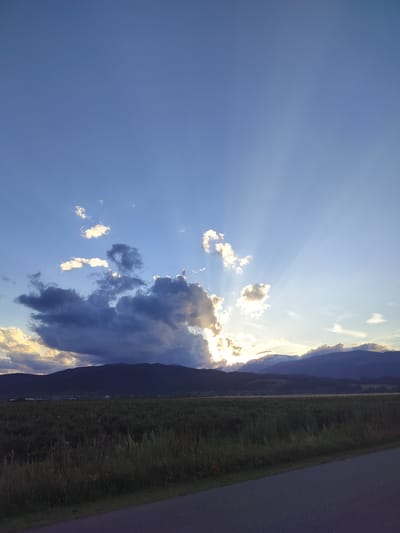 The image shows a wide shot of a rural landscape at sunset. The sky is a deep blue, with rays of sunlight emanating from behind dark clouds on the horizon. A range of silhouetted mountains forms the background, beyond which is a flat, dark field of crops. The foreground features a paved road running diagonally from the bottom right corner towards the left. The location is identified as Manolovo, Bulgaria. No people or specific actions are visible.