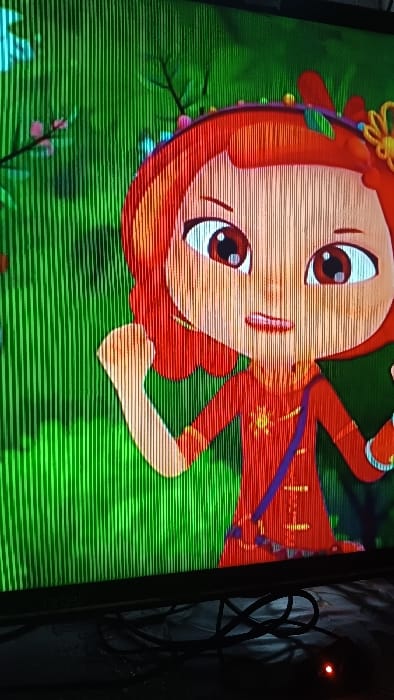 A close-up photograph taken in Kasansay, Uzbekistan, depicts an illuminated television screen displaying an animated scene. The screen exhibits prominent vertical scan lines across its surface. The central subject on the screen is an animated character resembling a young girl with reddish-orange hair, large brown eyes, and a smiling mouth with visible teeth. She wears a red garment adorned with a yellow sun-like emblem on the chest and a dark blue strap extending from her left shoulder. Her right arm is raised with a slightly clenched fist. Decorations, including what appear to be colorful beads and leaves, are present in her hair. The animated background is predominantly green, suggesting foliage or grass, with visible brown tree branches and small pink blossoms. Below the television screen, in the foreground, a dark surface supports several black electrical cables, some tangled. A small, red indicator light is visible on the lower right side of the foreground, suggesting an active electronic device. The area immediately surrounding the screen is dimly lit.