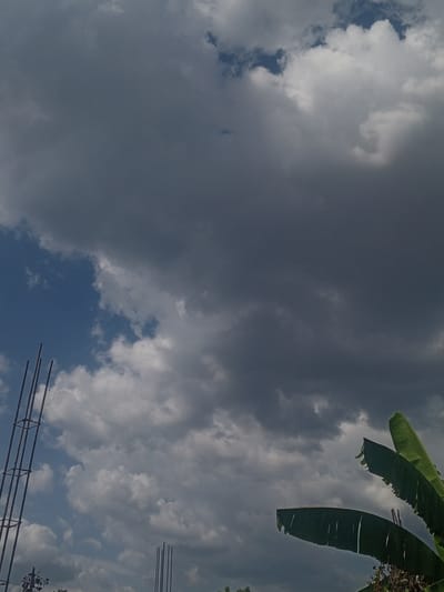 The image depicts a cloudy sky in Parroquia Ocumare, Venezuela. Dark, heavy storm clouds dominate the scene, contrasting with patches of blue sky. In the foreground, there are reinforced steel bars from a construction site and banana leaves. The overcast sky suggests impending rain or a recent storm.