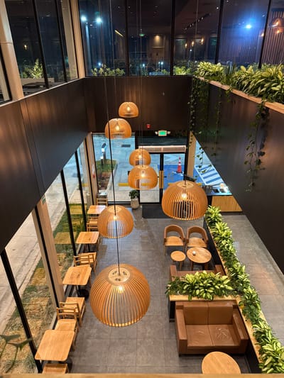 The image depicts the interior of a two-story establishment, likely a cafe or restaurant, located in Santa Tecla, El Salvador. The perspective is an overhead view from an upper level looking down into the main seating area. The scene is illuminated by artificial light, suggesting nighttime.
A prominent feature consists of seven large, spherical pendant lights with wooden slat designs, suspended at varying heights from the ceiling. The floor on the lower level is tiled in dark grey. Along the left side, adjacent to a floor-to-ceiling glass window wall, are multiple light-colored wooden square tables paired with matching wooden chairs, arranged for individual or small group seating. Beyond the window, an outdoor paved area with grass strips is visible.
On the right side, a lounge area features two brown leather sofas, several round wooden tables, and two individual light-colored wooden chairs. This area is bordered by a raised planter box filled with lush green foliage, including trailing vines that hang down the dark-colored wall. A small counter or bar area with bamboo-like paneling is partially visible in the back right.
At the rear of the lower level, a glass entrance/exit door leads to an exterior paved area, where a blue-painted section with white markings and an orange traffic cone are visible. An illuminated "EXIT" sign is mounted above this doorway. The upper level features additional glass window walls looking out onto an urban environment with illuminated foliage. No people are present in the scene.