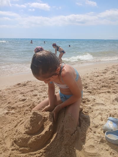 The media file shows a young girl playing in the sand on a beach. She is wearing a blue bikini and her hair is tied up. The girl is kneeling in the sand, with her back to the camera, and appears to be digging or building something. There are other people in the background, in the water, which suggests a warm, sunny day. The sea is calm and the sky is blue with some clouds. A pair of blue sandals are visible on the sand next to the girl. The scene is set in Kranevo, Bulgaria. The overall atmosphere is one of leisure and enjoyment at the beach.