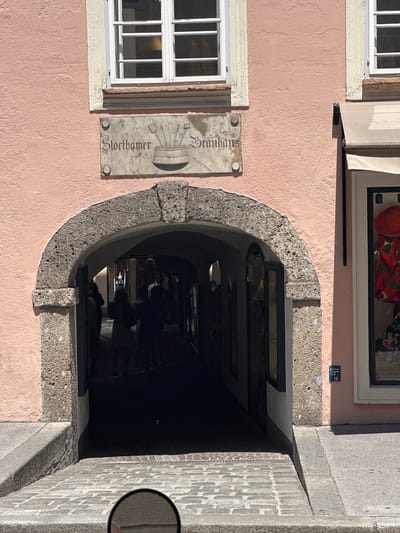 The image depicts an arched passageway leading into a building in Salzburg, Austria. The facade of the building is painted a pale pink. Above the archway, a stone plaque displays the inscription "Stortthamer Brauhaus" along with an image of a barrel and brewing tools. The arch itself is constructed of rough-hewn stone. Through the arch, a dimly lit interior arcade is visible, with several figures silhouetted within. To the right of the arch, a shop window displays colorful garments. The ground in front of the arch is paved with cobblestones. The overall scene captures a historical architectural detail in a European city.