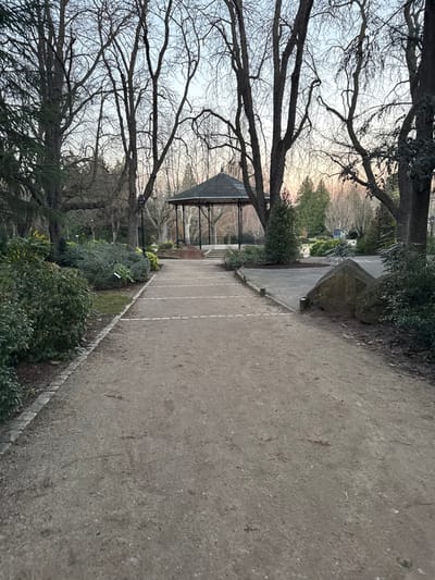 A long, winding dirt path stretches into the distance, bordered by manicured greenery on one side and towering, bare trees on the other. A gazebo sits midway along the path, partially obscured by tree branches. A large, angular rock formation stands near the path's edge. The scene suggests a peaceful park setting during the transition between seasons.