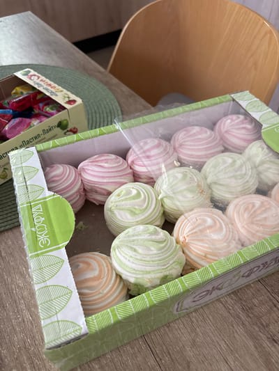 A close-up, eye-level shot captures a box of colorful marshmallows and candies on a wooden table, partially obscured by a green placemat. The box of marshmallows, made of clear plastic, reveals a variety of swirled marshmallows in pink, green, and orange hues. The packaging of the marshmallows features green, leaf-like designs and text that appears to be in Cyrillic script. To the left of the marshmallow box, a portion of a smaller cardboard box containing individually wrapped candies is visible. These candies are brightly colored in red, yellow, and pink wrappers. The background shows a light wooden chair and a subtle blur of the surrounding environment, suggesting an indoor setting. The lighting is natural and appears to be coming from the right side of the frame. The image is sharp and in focus, highlighting the textures and details of the confectionery items. The location of Vardane-Verino, Russia is not depicted or implied by any visual cues in the image.