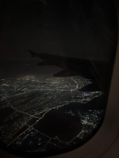 The image is an aerial view from an airplane window at night, overlooking a city illuminated by countless lights. A portion of the airplane's wing is visible in the upper right and upper left of the frame, silhouetted against the dark sky. The city below appears to be a sprawling urban landscape with a dense network of streets and buildings visible through the glowing lights. Roads can be traced as brighter lines within the darkness. In the distance, there are patches of haze or clouds that diffuse the city lights. There is a very faint light visible in the far upper portion of the image, possibly another aircraft. The image does not contain any discernible people or specific identifiable landmarks that would allow for a precise location within Zharmukhabmet, Kazakhstan. The scene is entirely urban and nocturnal.