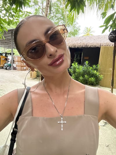 The media file shows a close-up, overhead shot of a woman posing outdoors. She is wearing large, light-brown sunglasses, glossy pink lipstick, and a beige top. A silver necklace with a cross pendant rests on her décolletage. She also has a large hoop earring in her visible ear. Her skin is visible, showing some freckles.

The background suggests an outdoor, tropical setting. Lush green foliage is visible in the foreground and behind her. In the distance, there appears to be a building with a thatched roof and bamboo walls, and some stacked boxes are visible. A small yellow piece of construction equipment is also in the background. The lighting suggests it is daytime, possibly overcast. The overall mood appears casual and relaxed. The woman's pose and direct gaze into the camera suggest she is taking a selfie or being photographed. There is no visible text in the image.