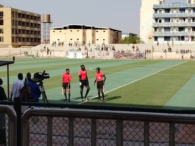 The image shows a soccer field in Bahri, Sudan. Three referees in red shirts and black shorts stand on the green and light green turf. A cameraman with a camera on a tripod stands to their left. A fence is in the foreground. In the background, spectators are seated along the top of the stadium wall, and banners with portraits adorn the wall. Buildings of light-colored brick are visible behind the wall, along with a water tower on the left side.