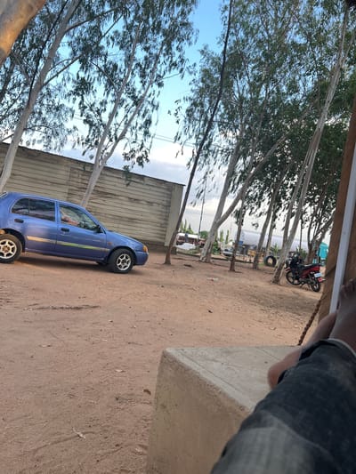 In Mayo Dasa, Nigeria, an outdoor scene unfolds with a clear sky and sparse trees. A small blue car is parked on a dirt lot, its side facing the viewer. Behind the car, a corrugated metal building stretches across the frame. In the distance, various vehicles are visible, including a white tanker truck, a red motorcycle, and other trucks. The foreground shows a person's legs and feet, clad in denim jeans, resting on a concrete structure. The ground is covered in dirt and scattered leaves, with a plastic bottle lying near the car. The overall impression is one of a rustic, functional area, possibly a roadside stop or a rural commercial space.