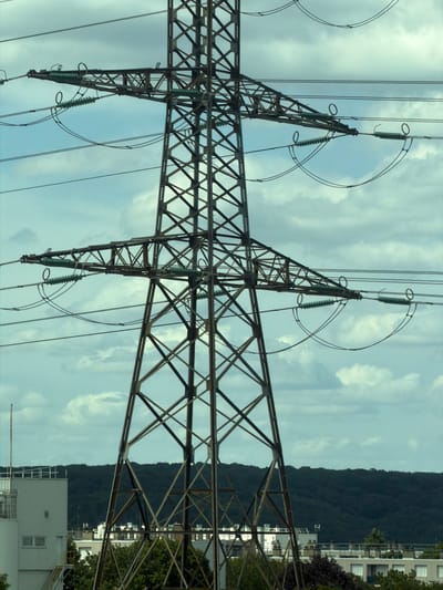 The image shows a large, lattice steel electricity pylon supporting power lines against a cloudy sky. The pylon is intricate in its construction, with many diagonal and horizontal crossbeams forming a strong, geometric structure. Several insulators, likely made of glass or ceramic, are visible, attached to the pylon and suspending the thick electrical cables. These cables stretch across the frame, some arcing slightly.

In the background, a dark, forested hill rises steeply, indicating a natural landscape. To the right of the hill, several modern buildings are visible, suggesting a developed area adjacent to the natural environment. These buildings have a clean, functional design with windows and balconies. The sky is filled with a mix of white and gray clouds, suggesting overcast or partly cloudy weather. The overall lighting is diffused, which could imply it is daytime, but the specific time is not clearly discernible.

The image appears to be taken from a low angle, looking up at the towering pylon and the sky. The composition emphasizes the scale and industrial nature of the pylon in contrast with the natural and built environments in the distance. There is no visible text in the image. The scene is devoid of people, focusing solely on the infrastructure and the surrounding landscape. The orientation of the image is such that the ground appears to be at the top of the frame, and the sky at the bottom, suggesting it may have been rotated.