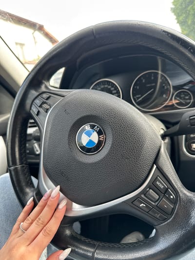 The image shows the interior of a car, specifically a BMW steering wheel. The center of the steering wheel prominently displays the BMW logo. To the right of the logo are buttons and controls. Behind the steering wheel, the car's gauges are visible. A woman's hand with long nails is on the lower right portion of the steering wheel. A silver ring is visible on her ring finger. The location in the city of Djerman, Bulgaria is unknown.