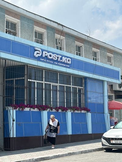 In Kadamjay, Kyrgyzstan, an elderly woman with short, gray hair, wearing a black and white tunic over a black skirt and white sandals, walks along a sidewalk in front of a blue building. She carries a dark bag in her right hand. The building has a prominent blue facade with the "POST.KG" logo and Cyrillic text. Numerous windows with metal grates are visible on the upper floors, and planters filled with purple flowers line the lower section of the building. To the right of the building, a portion of a white car with a license plate reading "G3 532 AGR" is parked on the street. A red umbrella is partially visible behind the car. The environment appears to be an urban setting with a clear sky.