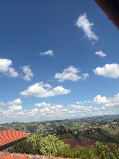 From an elevated perspective, the photo captures a broad landscape punctuated by a bright blue sky dotted with scattered cumulus clouds. In the lower portion, the scene transitions to the urban environment of Castiglione Tinella, Italy, where vineyards are visible between buildings with red-tiled roofs, some of which are partially obscured by lush green foliage. The focus of the photograph is on the interplay between the natural elements and the built environment, showcasing the rural aspects of the setting. No people are visible in the image.