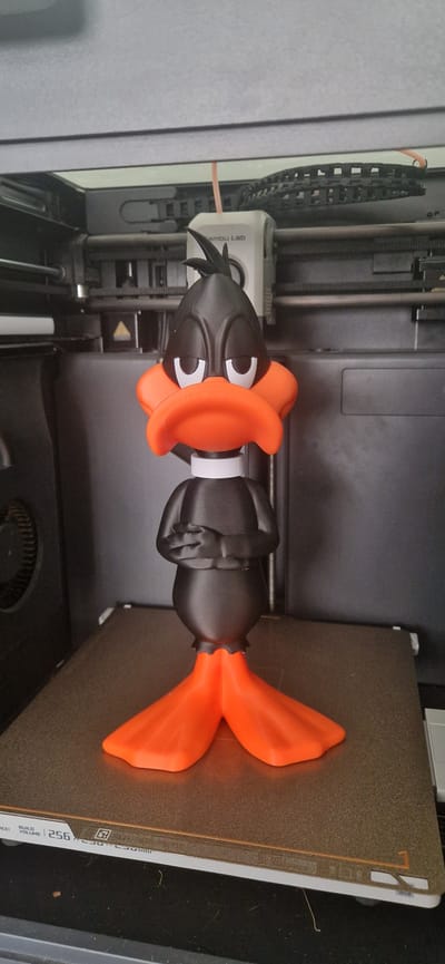 A 3D printed Daffy Duck figure stands on a textured build plate of a 3D printer. The figure is black with an orange beak and feet, and a white collar. Daffy Duck's arms are crossed, and its expression is apathetic. The 3D printer is open, revealing its internal components, including a white print head with the "Bambu Lab" logo visible. The environment appears to be indoors, likely a workshop or a room within a residential property. The image does not provide any information about the city of South Holland, United Kingdom, as the context is a domestic 3D printing setup.