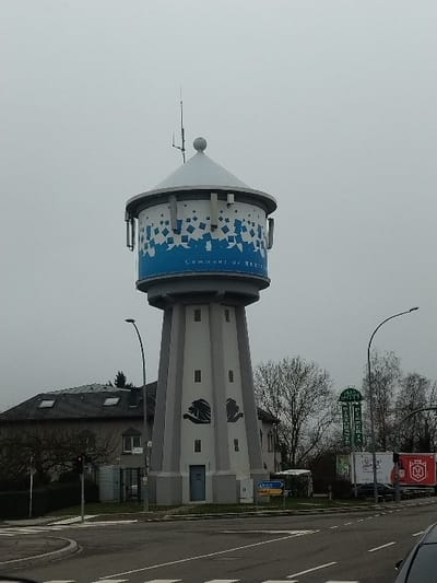 A large, multi-faceted water tower dominates the center of the image, located at an intersection in Bertrange, Luxembourg, as indicated by "COMMUNE DE BERTRANGE" written on its upper blue band. The tower features a light grey, tapering base with a light blue door and two dark silhouetted lion head motifs. The cylindrical top section has a blue band with white geometric patterns, supporting a white conical roof topped with a finial and multiple communication antennas. Several rectangular grey communication units are affixed to the circumference of the cylindrical section.

To the left of the water tower, a two-story building with a dark grey hip roof and multiple skylights is visible, partially obscured by bare trees. A red traffic light is active at the intersection in the foreground.

To the right of the water tower, a tall green sign for "PIZZERIA RISTORANTE" is visible, with text including "IL GIARDINO" and "SAINT NICOLAS." Behind this sign, two white commercial vehicles or containers are parked, one displaying a red and white logo. Bare deciduous trees are scattered throughout the background.

The foreground consists of an asphalt road with white lane markings, including a crosswalk and a stop line. A blue and yellow directional sign is located near the base of the water tower, partially displaying "AUBANGE" and "COMMU." Another small sign with the number "11" is also visible. The sky is uniformly overcast and light grey, suggesting a dull, daytime environment. No individuals are present in the visible scene.