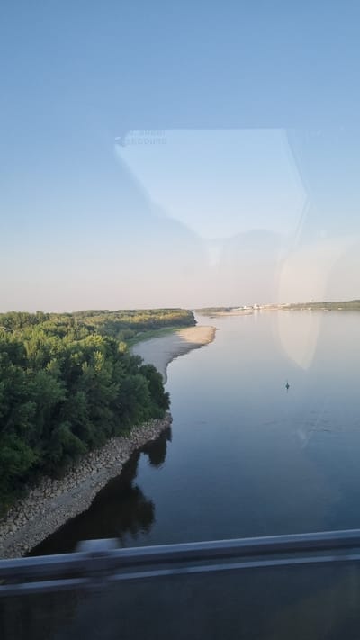 The image shows a wide river with a tree-lined bank on the left. A sandy strip is visible along the riverbank, and a few buildings can be seen in the distance on the opposite side of the river. A green buoy floats in the water. The sky is clear and blue, with some faint reflections on a glass surface in the foreground. The scene appears to be a natural landscape with some man-made structures in the distance.