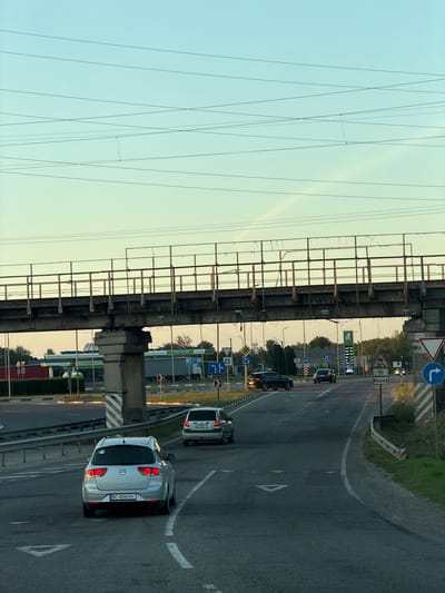 This media file depicts a road scene in Stryi, Ukraine. The perspective is from a moving vehicle, looking towards the right side of the road. The sky is a soft gradient of pale blue and yellow, suggesting it is either early morning or late evening.
The scene features a paved road with multiple lanes, marked by white lines. Several cars are visible on the road, including a silver car in the foreground with license plate BC 4044 HI, and a white car further ahead. A large concrete structure, possibly part of a bridge or overpass, runs along the left side of the road. Overhead, a complex network of wires stretches across the sky.
Along the right side of the road, there are various road signs. One sign indicates a turn to the left, another displays a warning about slippery roads. A sign with a snowflake and "6км" suggests a distance to a location. There is also a sign indicating a parking area and a gas station with prices displayed on a digital screen. Green bushes and trees line the edge of the road in the background. The overall impression is of a typical highway scene during daylight, with evidence of infrastructure and traffic.
