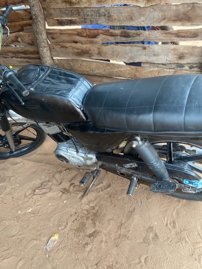 A black motorcycle is parked on sandy ground in Okura, Nigeria. The motorcycle is positioned adjacent to a wooden structure. Part of a person wearing a blue shirt is visible through the wooden structure. A piece of plastic is lying on the sand.