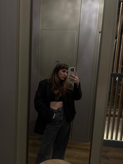 A woman takes a mirror selfie indoors, likely in a fitting room or bathroom due to the presence of a toilet sign on the wall. She is wearing a black blazer over a black crop top and distressed denim jeans. Her hair is long and brown with bangs. The environment appears to be modern, with neutral colored walls and wooden flooring. The background also shows a glimpse of an outdoor area with vertical wooden slats and some greenery.